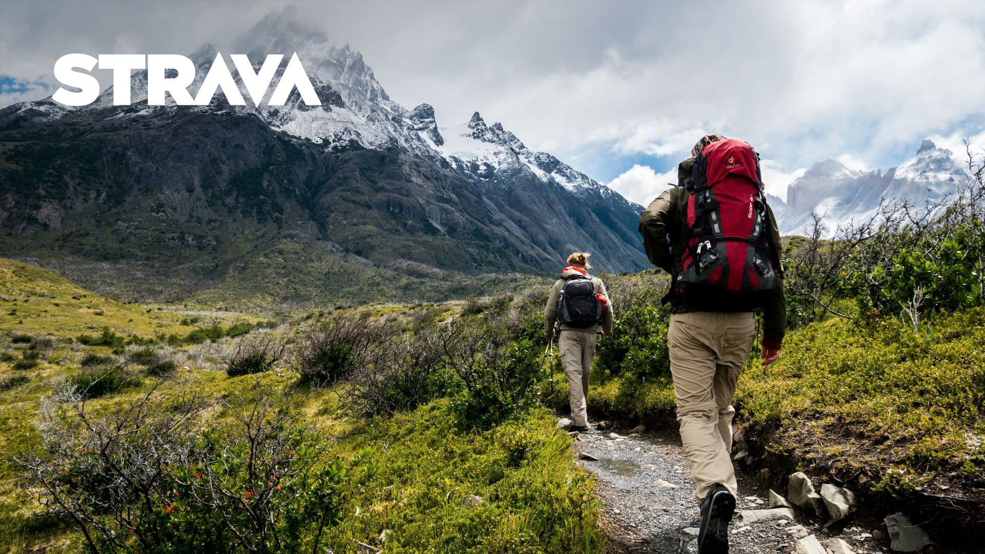 Two people backpack through an alpine environment with snowcapped mountains in the background